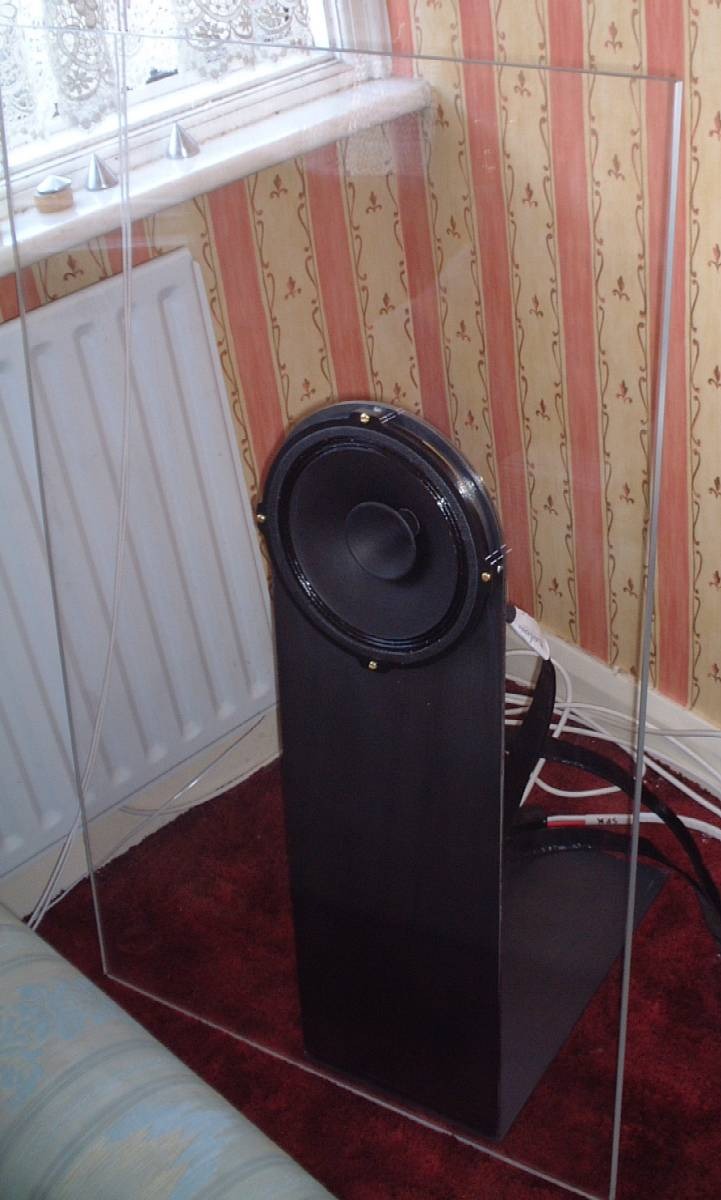 Photograph: the clear acrylic baffle in front of a black stand with the driver, in a corner of the room